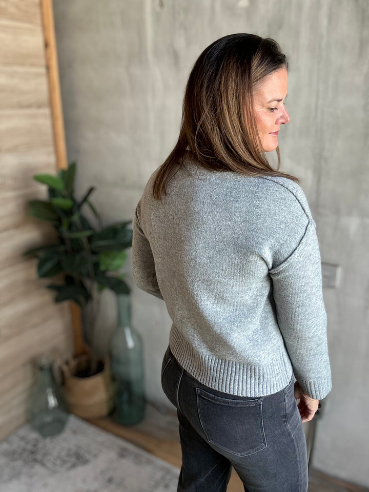 A woman wearing a light gray knit sweater and dark gray jeans, photographed from the back in an indoor setting with a concrete wall, wooden paneling, and potted plants.