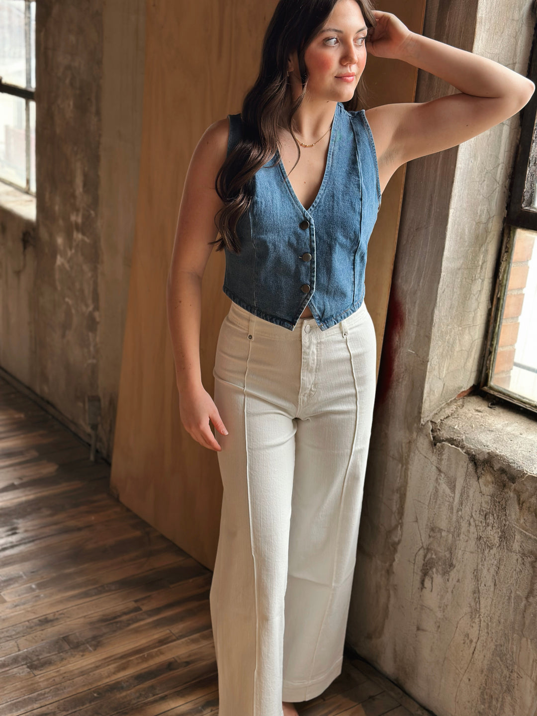 Woman wearing a denim vest and white pants standing in a room with wooden floor and stone walls.