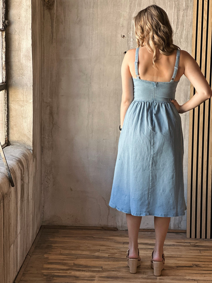 Back view of model wearing a flare dress that has a stretch denim fit with a sweetheart neckline, wide straps, and it is a light Denim color 