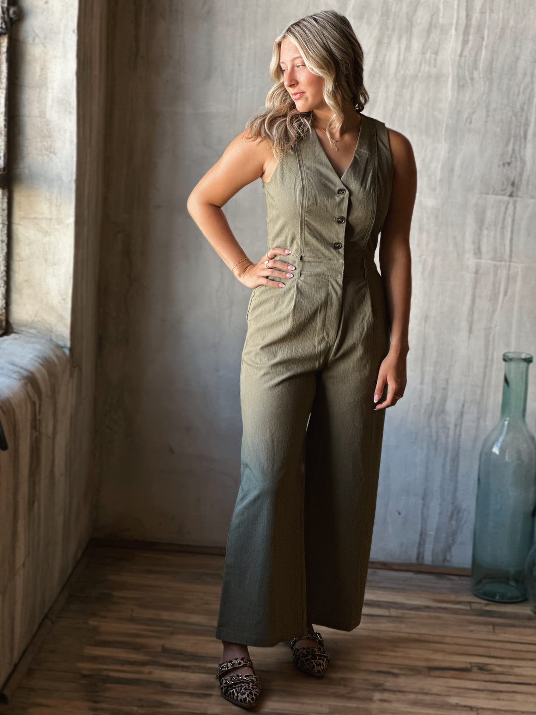 Woman wearing a sleeveless top and wide-leg pants in a room with wooden floor and textured wall.