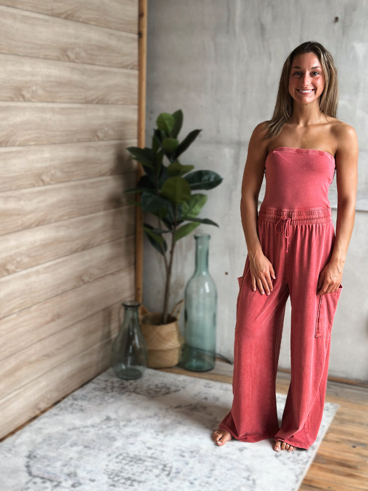 Woman wearing a light washed red strapless jumpsuit standing in a room with a plant and decorative bottles.