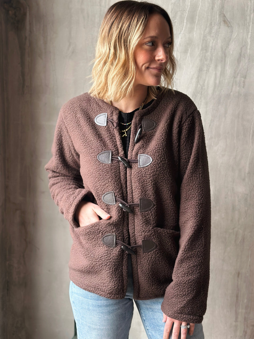 Woman wearing a brown fleece jacket with silver buttons against a gray background