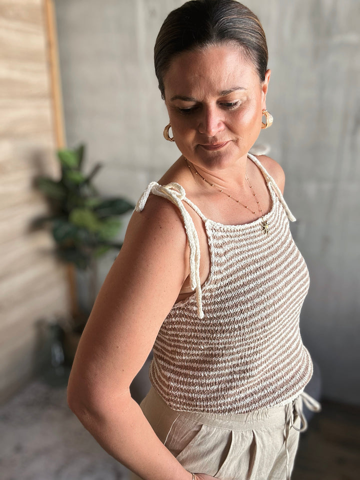 A woman wearing a light brown and beige striped crocheted tank top with thin straps that are tied into small bows.