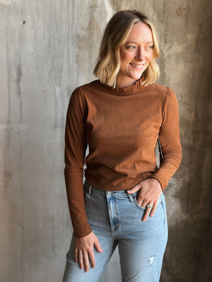 Woman wearing a brown long-sleeve top and light blue jeans against a concrete wall.
