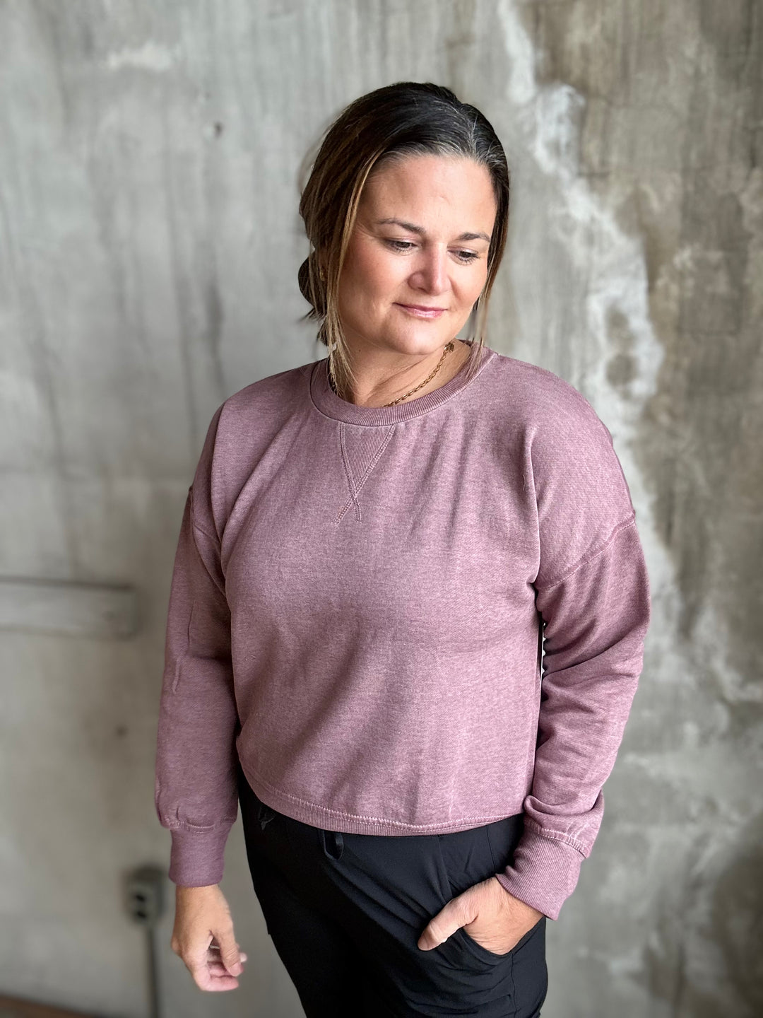 A woman wearing a dusty rose long-sleeve crewneck sweatshirt and black pants, standing against a textured concrete wall.