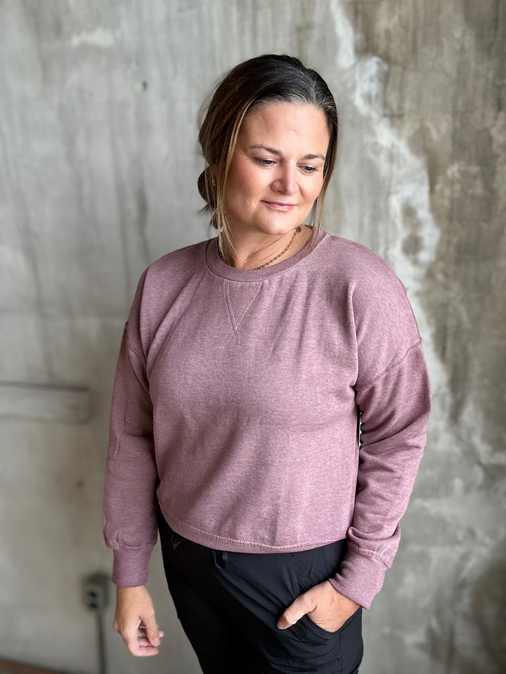 A woman wearing a dusty rose long-sleeve crewneck sweatshirt and black pants, standing against a textured concrete wall.