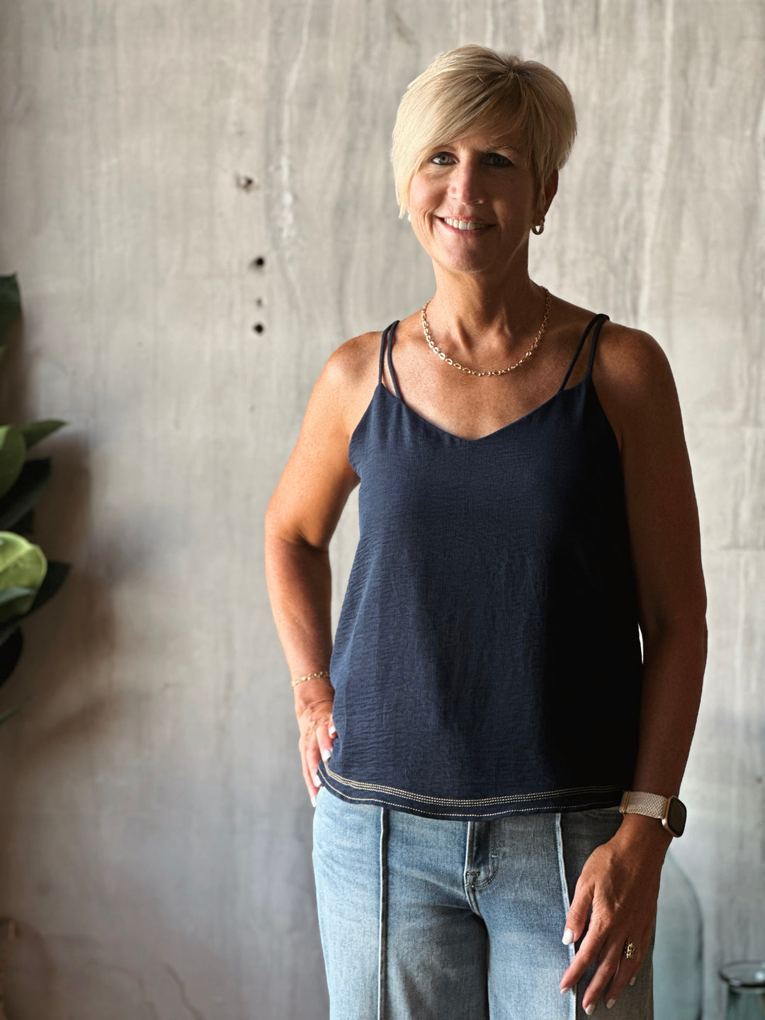 Woman wearing a navy tank top and light blue jeans standing against a textured wall.