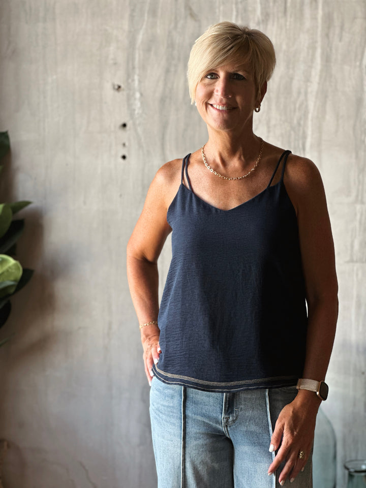 Woman wearing a navy tank top and light blue jeans standing against a textured wall.