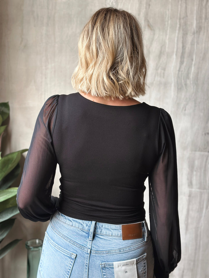 Person wearing a black top with sheer sleeves and light blue jeans against a neutral background