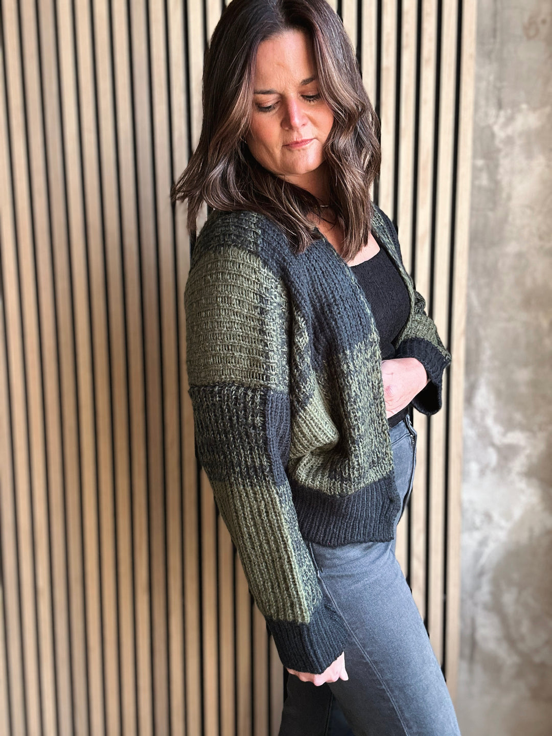Woman wearing a green and gray cardigan against a wooden paneled wall.
