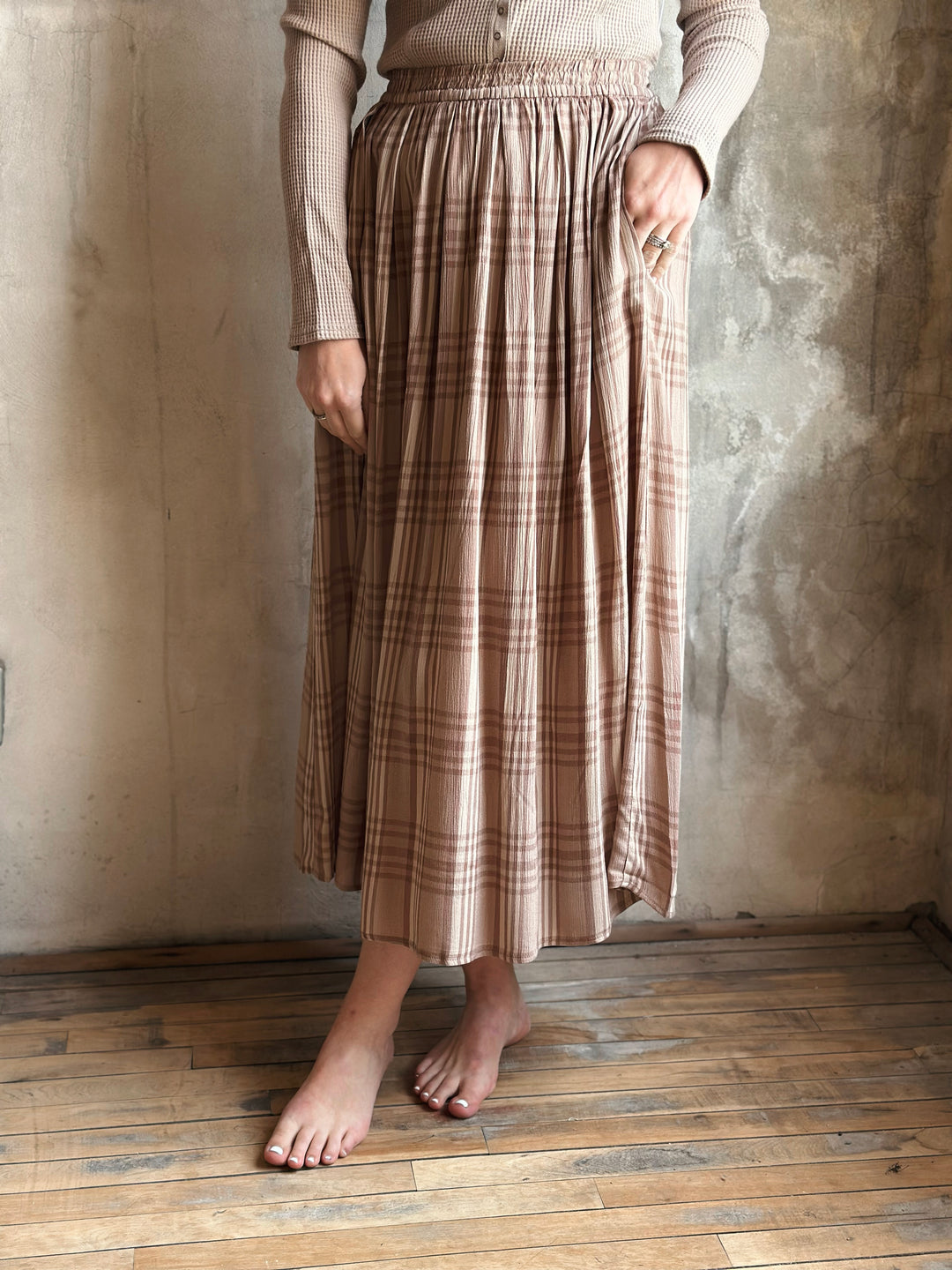 Person wearing a plaid skirt and beige top against a textured wall.
