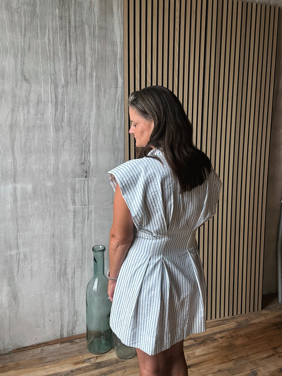 Woman in a striped dress standing against a textured wall with a vase on the floor.