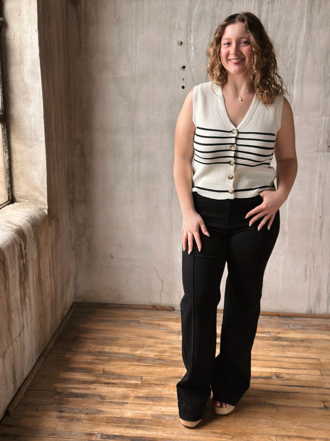 Woman wearing a white vest with black stripes and black jeans standing in a room with wooden floor and stone wall.