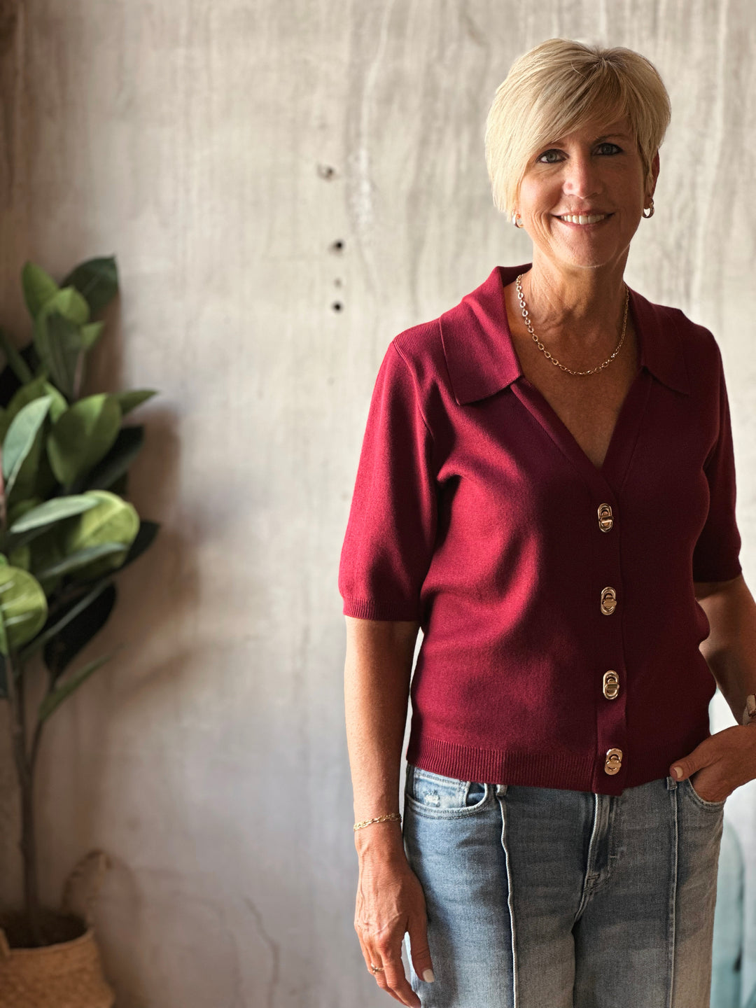 Woman wearing a red cardigan with gold buttons standing indoors next to a plant.