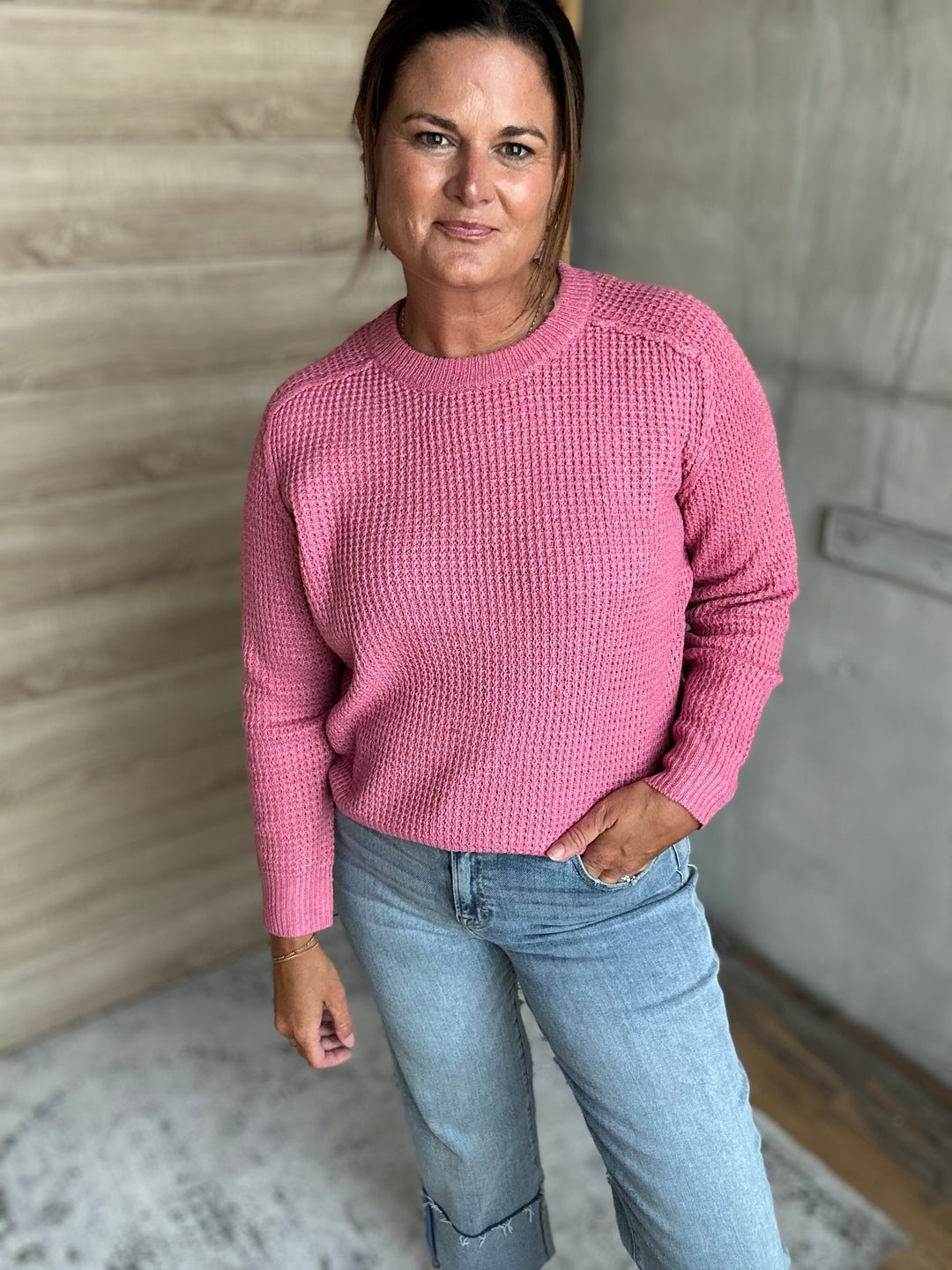 A woman wearing a textured pink long-sleeve knit sweater and light blue jeans, standing in an indoor setting with a wood-paneled wall and concrete wall in the background.