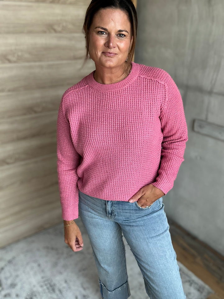 A woman wearing a textured pink long-sleeve knit sweater and light blue jeans, standing in an indoor setting with a wood-paneled wall and concrete wall in the background.