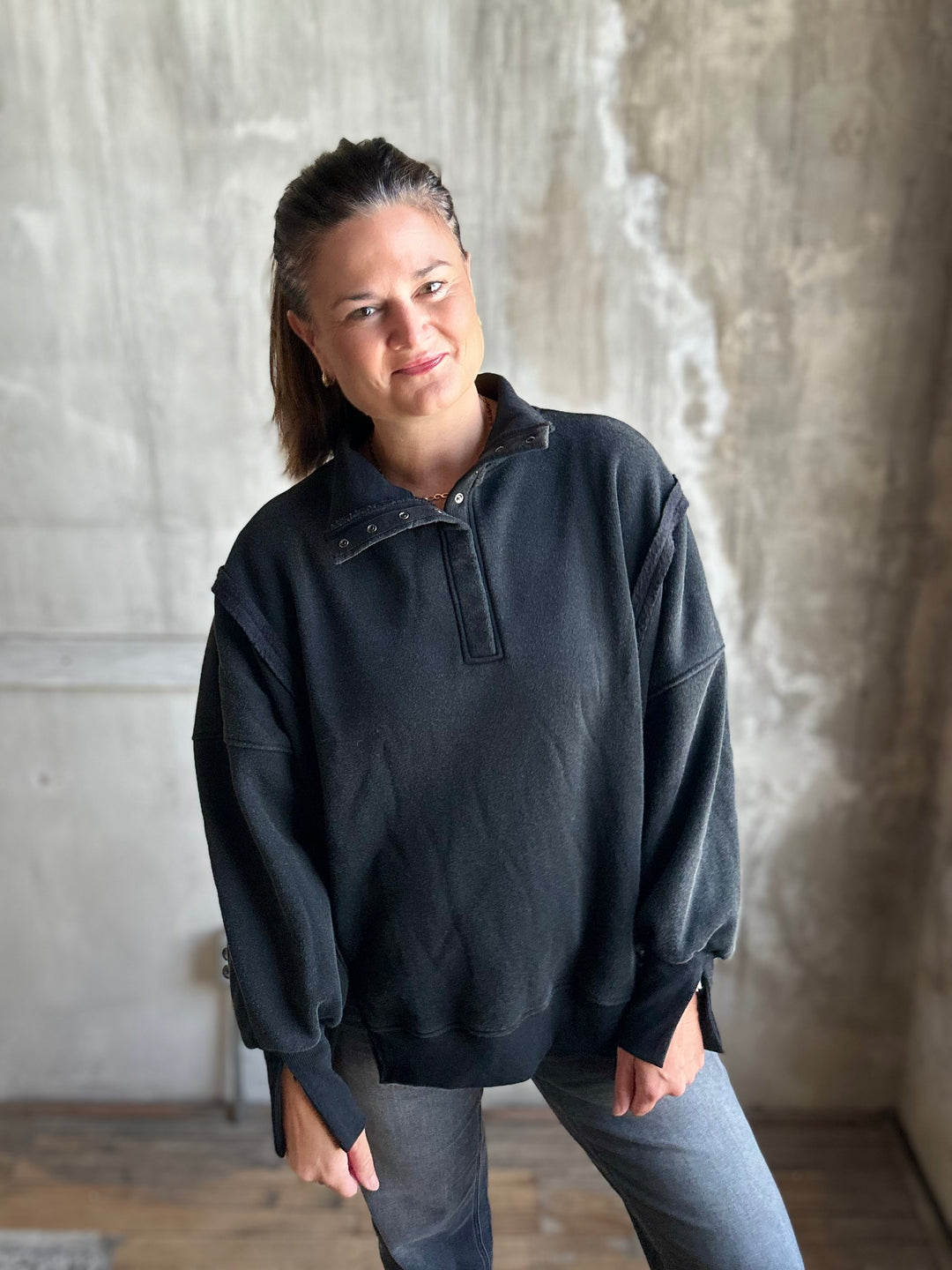 A woman wearing a dark charcoal gray half-placket pullover sweatshirt and gray jeans, standing against a textured concrete wall.
