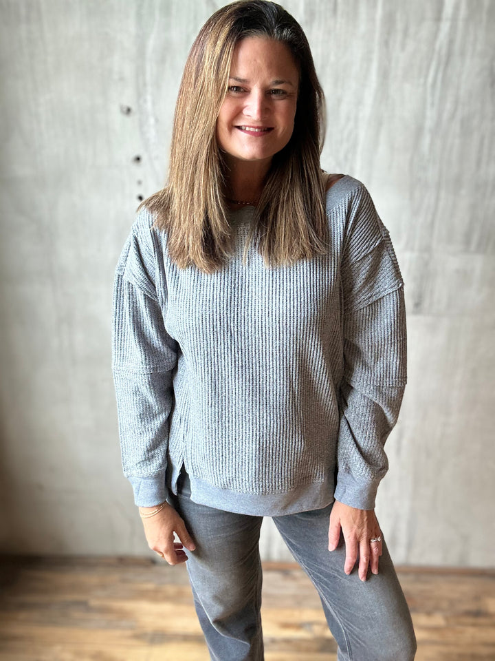 A woman with shoulder-length brown hair wearing a light gray ribbed knit long-sleeve sweater and gray pants, standing against a textured gray concrete wall with a wooden floor.