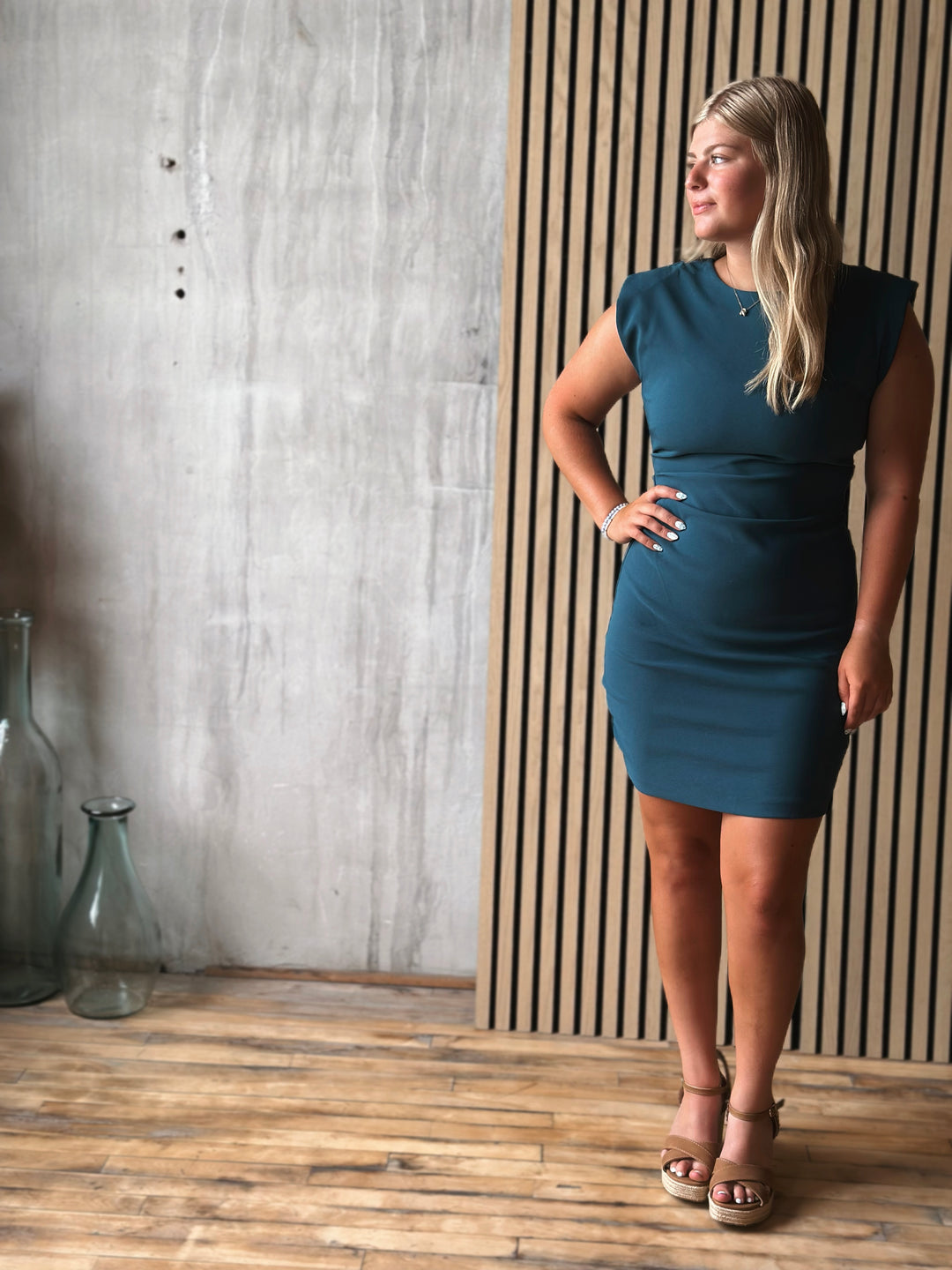Woman in a teal dress standing against a wooden paneled wall.