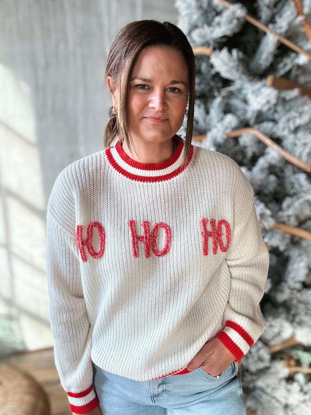 Person wearing a white sweater with 'HO HO HO' in red text, standing in front of a decorated Christmas tree.