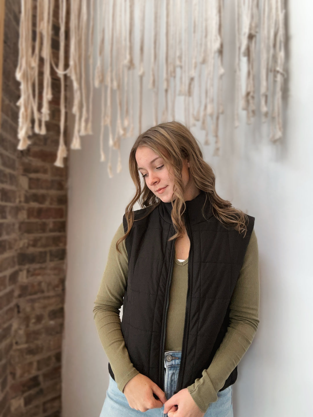 Woman wearing a black vest over an olive green long-sleeve shirt against a wall with hanging macrame art.