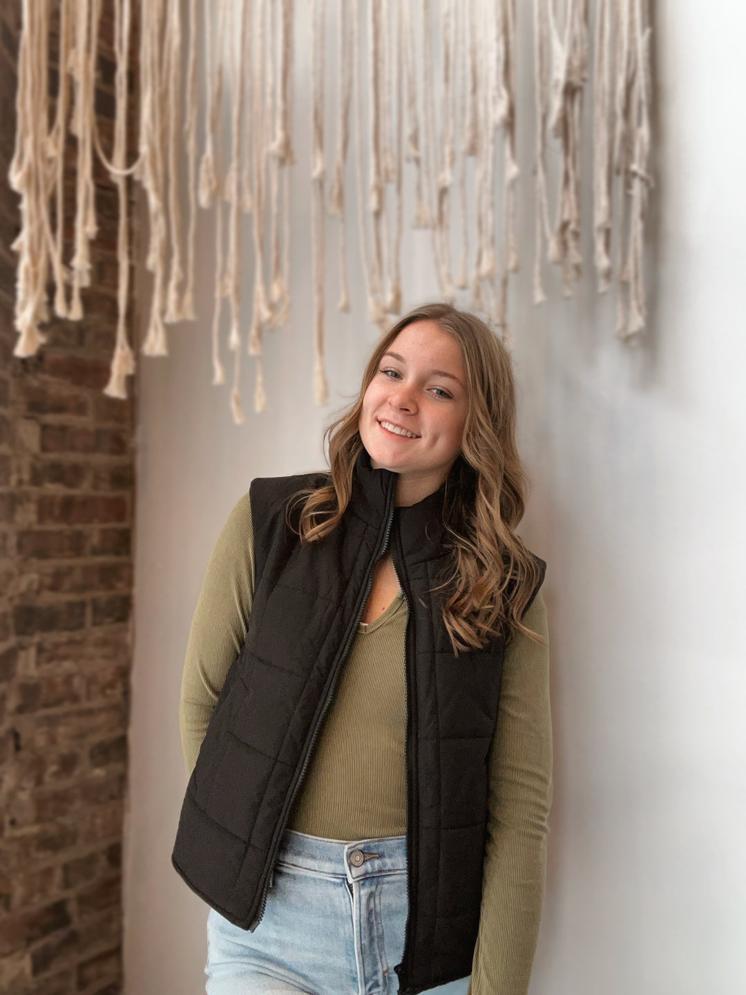 Woman wearing a black vest and green sweater against a wall with hanging fabric.