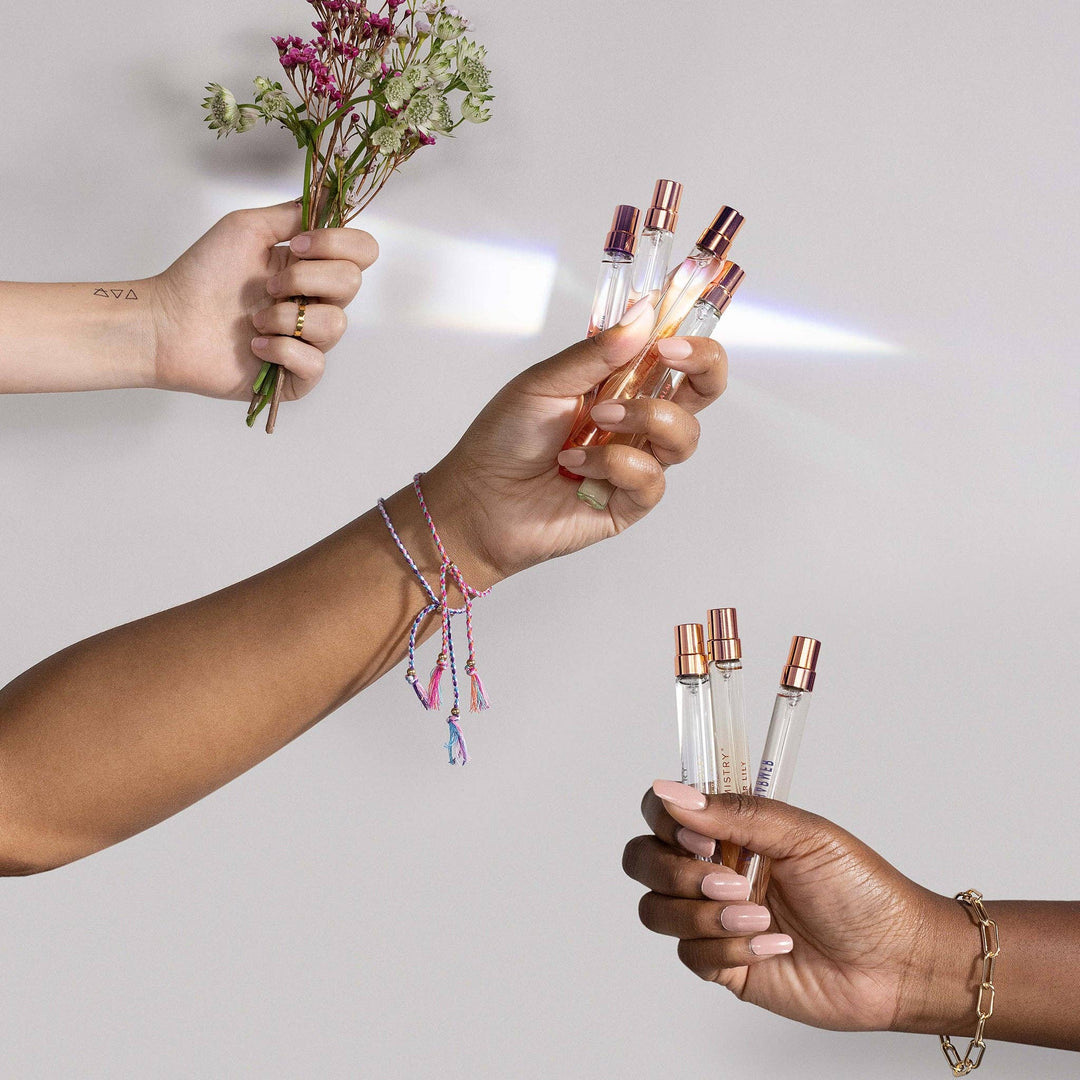 Three hands holding small perfume bottles and a bouquet of flowers against a plain background