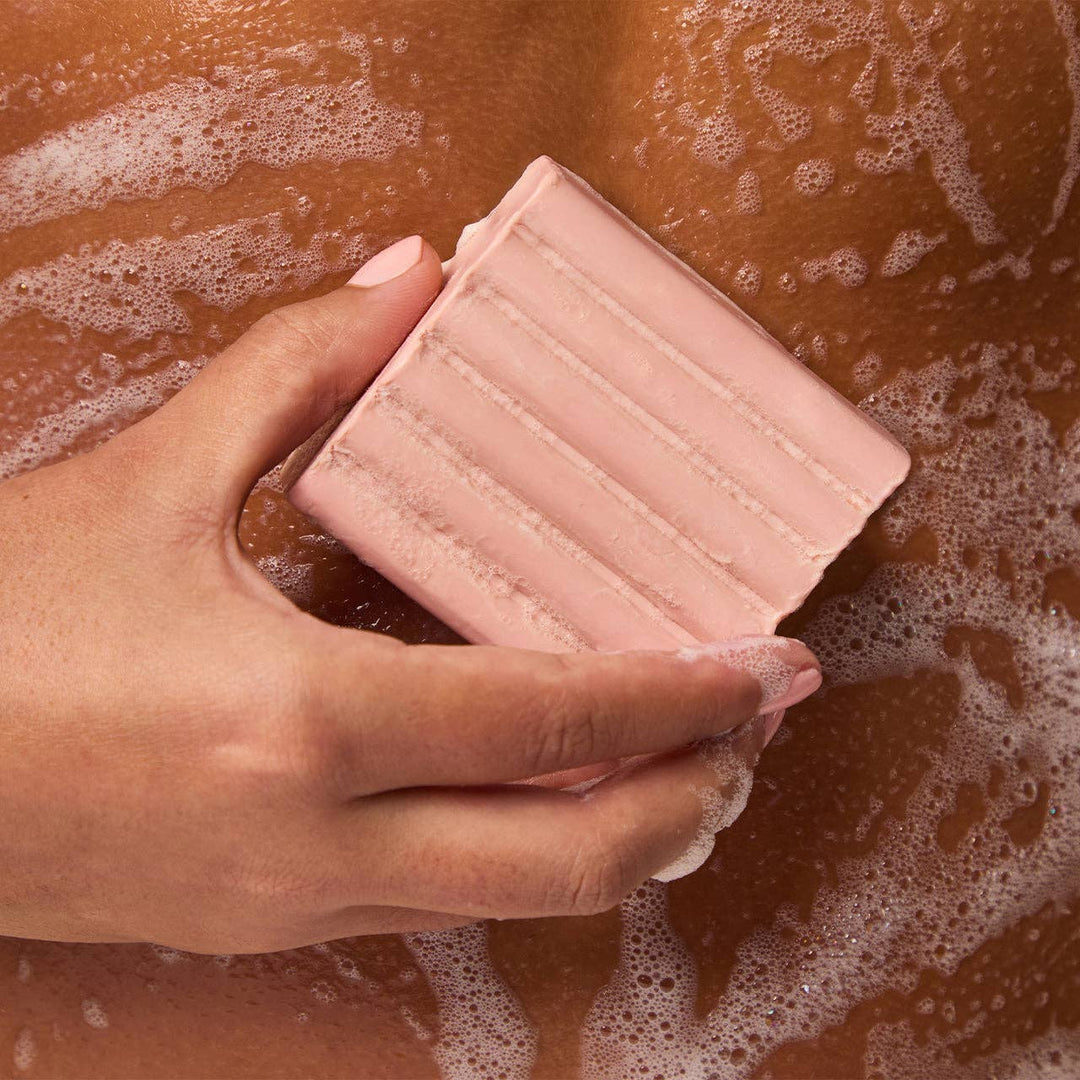 Hand holding a pink bar of soap with soap suds in the background