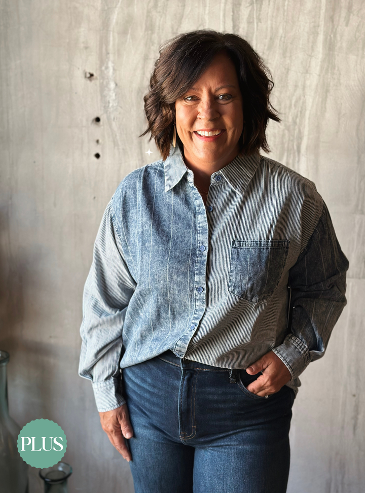 Woman wearing a light blue denim shirt and dark jeans against a concrete wall.